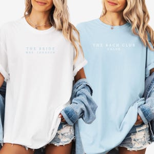 Op de afbeelding: Twee vrouwen dragen witte en lichtblauwe T-shirts. Het witte shirt heeft de tekst "THE BRIDE MRS. JOHNSON" in lichtblauwe tekst. Het lichtblauwe shirt heeft de tekst "THE BACH CLUB CHLOE" in witte tekst. Beide shirts hebben opgerolde mouwen.