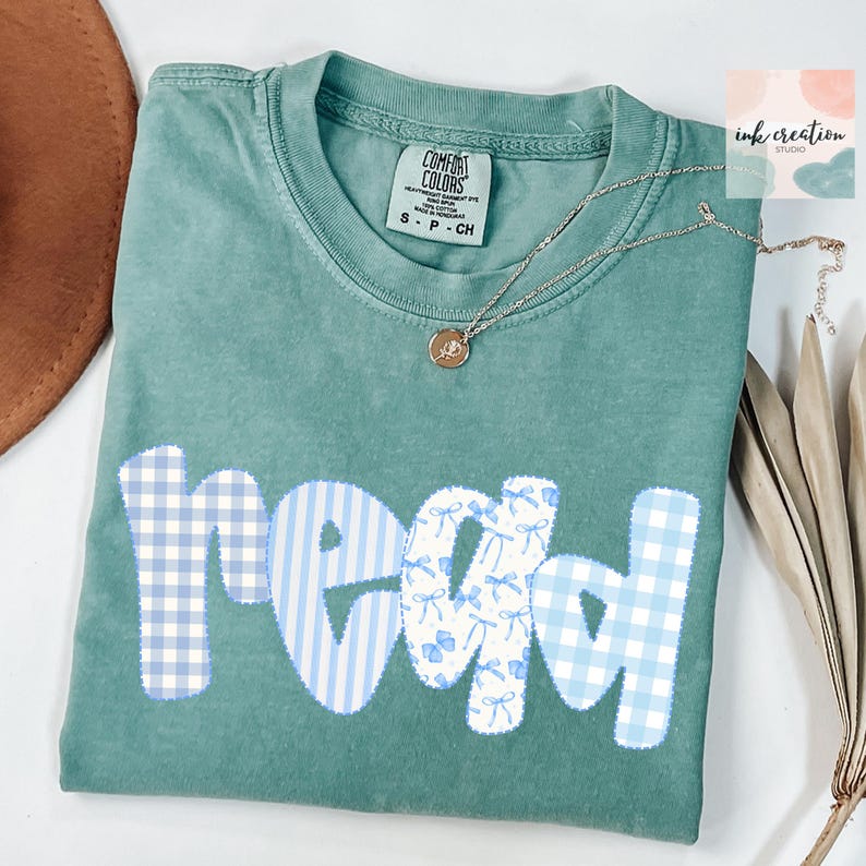 以下が含まれることがあります： 水色のComfort Colors Tシャツ。 「read」の文字は、青いギンガムチェックとリボンの模様を組み合わせています。 ペンダント付きのシルバーネックレスが折りたたまれたシャツの上にかけられています。