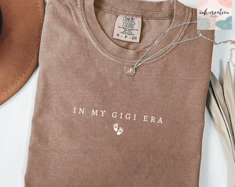 May include: A brown Comfort Colors t-shirt with the phrase "IN MY GIGI ERA" and baby footprints printed in white. A silver necklace with a pendant is draped across the shirt. A brown hat is partially visible in the upper left corner.