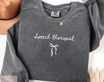 May include: A charcoal gray sweatshirt with the words "Speech therapist" embroidered in white script, along with a white bow graphic. A gold necklace and a brown hat are visible in the background.