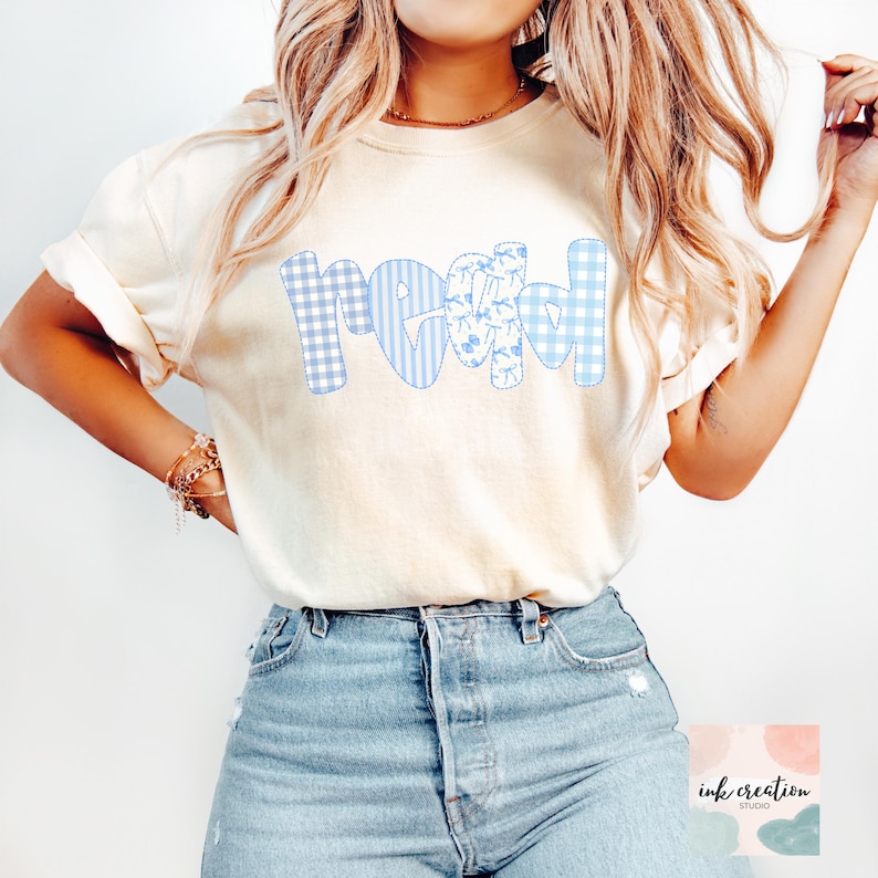 以下が含まれることがあります： クリーム色のTシャツ。青色のパッチワーク文字で「read」と書かれています。文字はギンガム、ストライプ、花柄です。モデルはブルージーンズを着用しています。画像には「ink creation studio」のロゴが含まれています。