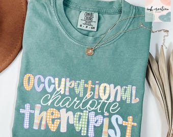 May include: A teal Comfort Colors t-shirt with the words "Occupational Charlotte Therapist" in a patchwork design. A silver necklace with a small pendant rests on the shirt. The shirt is folded, with a brown hat visible in the upper left corner.