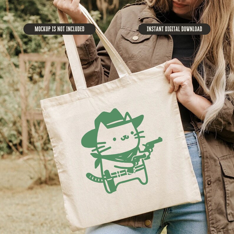 Op de afbeelding: Een witte tote bag met een groene cartoonkat die een cowboyhoed draagt en een pistool vasthoudt. De tekst "MOCKUP IS NOT INCLUDED" staat bovenaan de tas.
