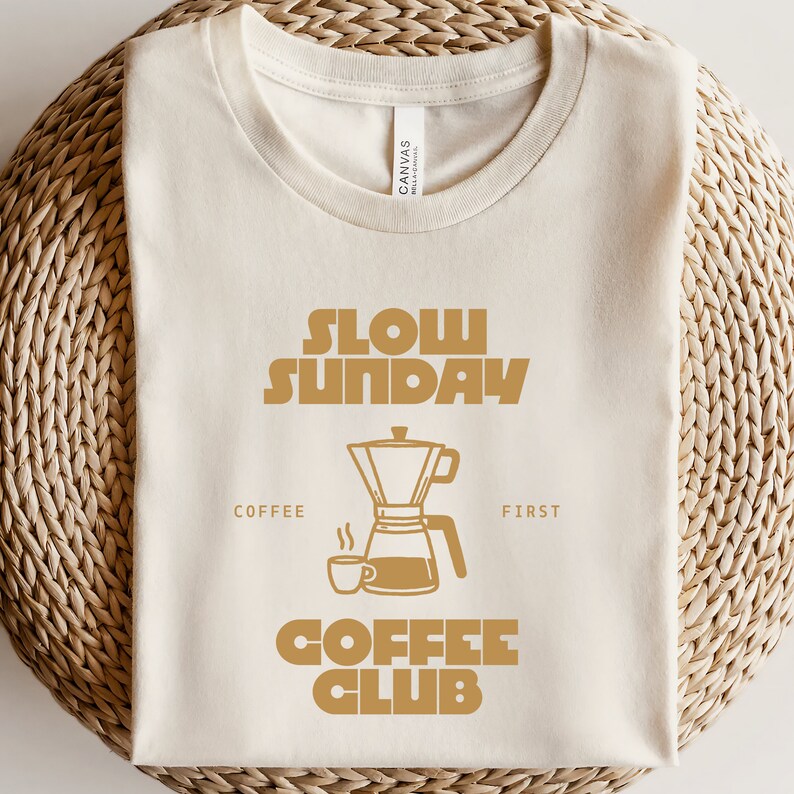 May include: A beige t-shirt with the text "Slow Sunday Coffee Club" in brown lettering. The design includes a coffee pot and a coffee cup.