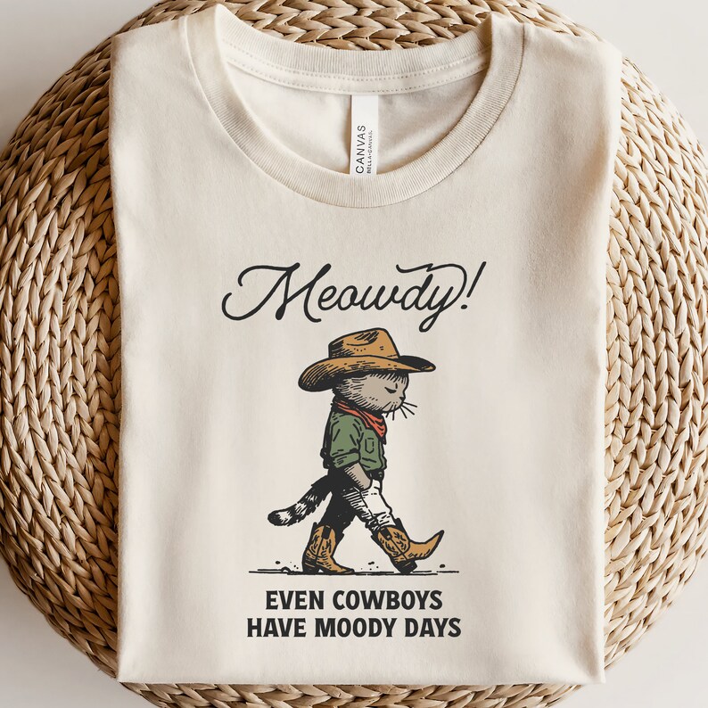 May include: A beige t-shirt featuring a cartoon illustration of a cat wearing a cowboy hat and boots. The text "Meowdy!" is above the cat and "Even cowboys have moody days" is below the cat.