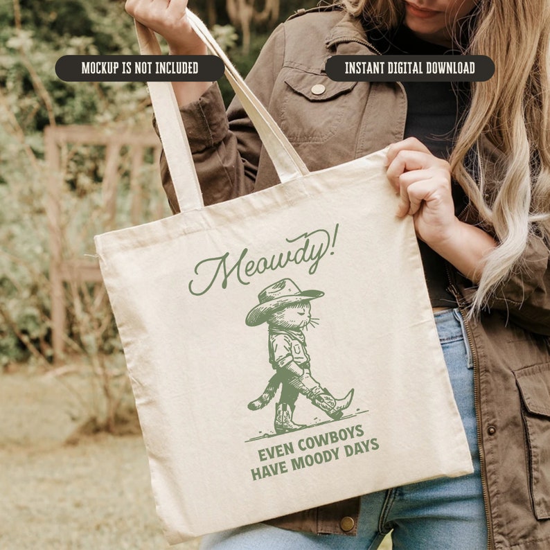 May include: A white tote bag with a green illustration of a cat wearing a cowboy hat and the text "Meowdy! Even cowboys have moody days".
