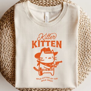 Op de afbeelding: Een beige T-shirt met een oranje afbeelding van een kat met een cowboyhoed en een pistool. De tekst "Killer Kitten" staat boven de kat en "Wild Kitten of the Wild West" staat onder de kat.