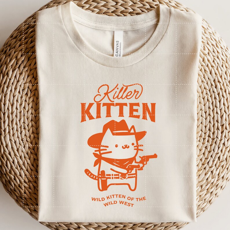 Op de afbeelding: Een beige T-shirt met een oranje afbeelding van een kat met een cowboyhoed en een pistool. De tekst "Killer Kitten" staat boven de kat en "Wild Kitten of the Wild West" staat onder de kat.