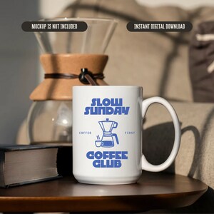 May include: White ceramic coffee mug with blue text that reads "Slow Sunday Coffee Club" and a blue illustration of a coffee pot. The mug is on a wooden table.