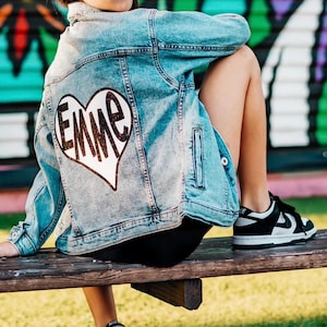 May include: A light blue denim jacket featuring a large white heart graphic on the back with the name EMME written in dark brown glittery letters, paired with black leggings and black and white low-top sneakers with a side swoosh logo.