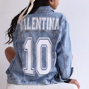 May include: A light blue acid-wash denim jacket featuring the name VALENTINA in white block letters across the upper back, with the large white number 10 printed below. The jacket has a classic collar, long sleeves, and visible seam detailing.