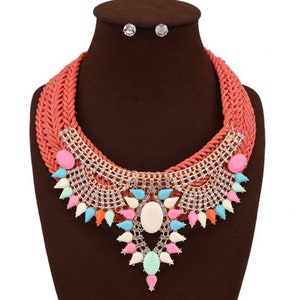 May include: A coral colored woven choker necklace with a large statement pendant featuring a white oval stone surrounded by smaller colorful stones and gold-toned metal accents.