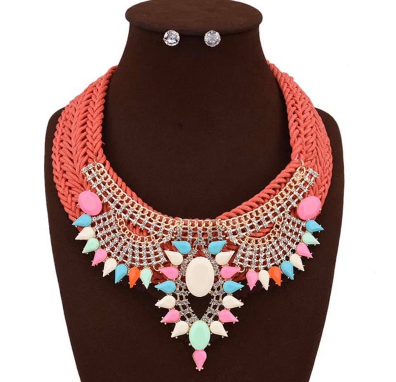 May include: A coral colored woven choker necklace with a large statement pendant featuring a white oval stone surrounded by colorful teardrop shaped stones and rhinestones. The necklace comes with a matching pair of earrings with clear rhinestones.