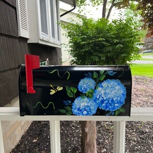 Puede incluir: Buz&oacute;n negro con un dise&ntilde;o pintado a mano de hortensias azules y hojas verdes. Una lib&eacute;lula amarilla est&aacute; pintada en la parte delantera del buz&oacute;n.