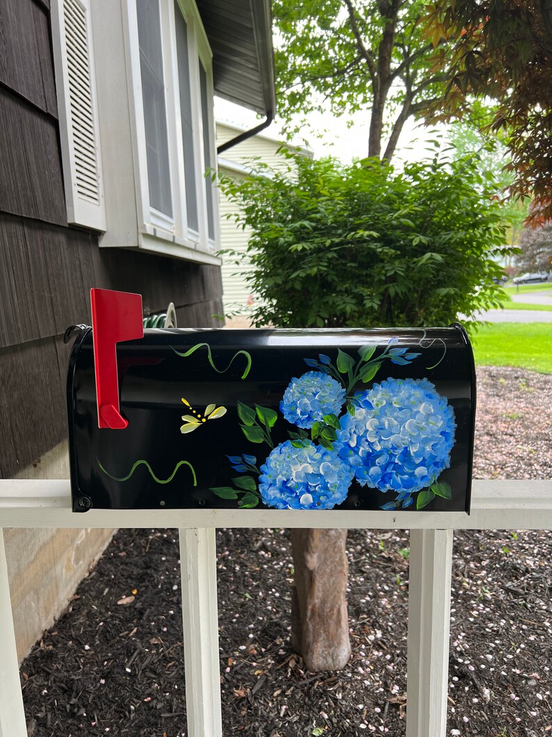 Puede incluir: Buz&oacute;n negro con un dise&ntilde;o pintado a mano de hortensias azules y hojas verdes. Una lib&eacute;lula amarilla est&aacute; pintada en la parte delantera del buz&oacute;n.