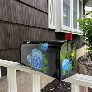 Puede incluir: Buz&oacute;n negro con un dise&ntilde;o pintado de hortensias azules y mariposas amarillas. El buz&oacute;n est&aacute; montado en una valla blanca de madera.