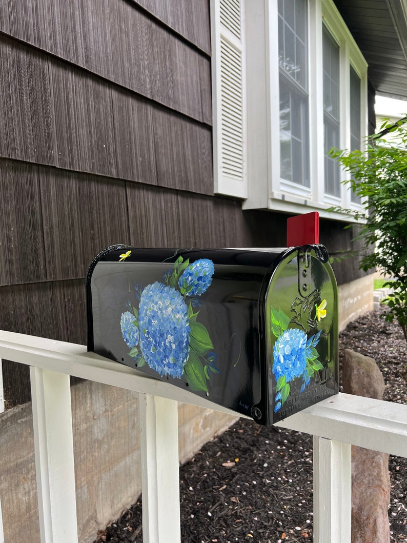 Puede incluir: Buz&oacute;n negro con un dise&ntilde;o pintado de hortensias azules y mariposas amarillas. El buz&oacute;n est&aacute; montado en una valla blanca de madera.