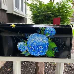 Puede incluir: Buz&oacute;n negro con un dise&ntilde;o pintado a mano de hortensias azules y hojas verdes. El buz&oacute;n est&aacute; montado en una valla blanca de madera.