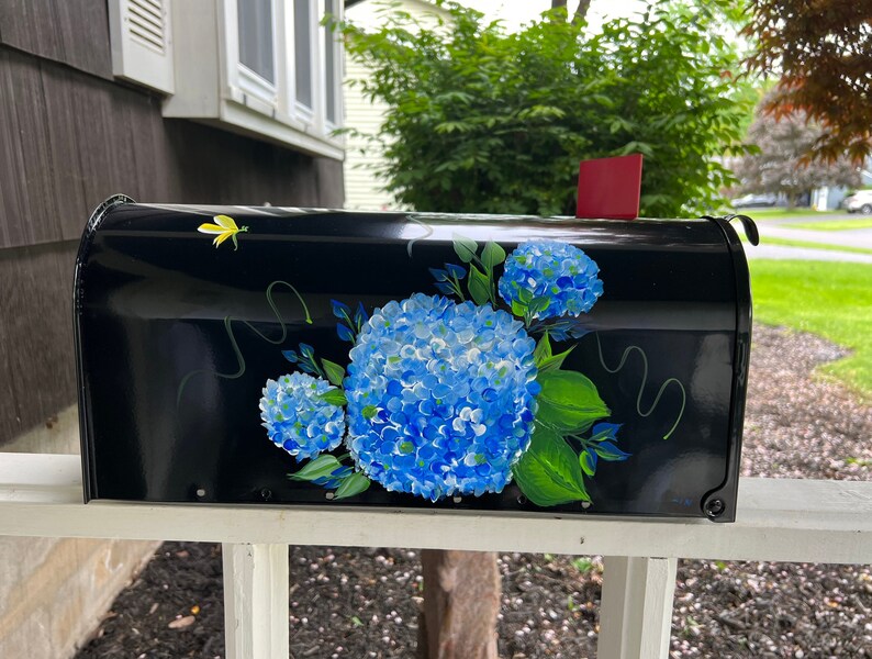 Puede incluir: Buz&oacute;n negro con un dise&ntilde;o pintado a mano de hortensias azules y hojas verdes. El buz&oacute;n est&aacute; montado en una valla blanca de madera.