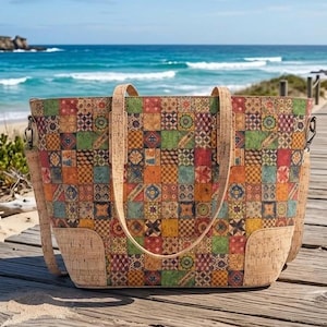 May include: A colorful tote bag with a patchwork design. The bag features a variety of square patterns in shades of red, orange, green, and blue. The handles and base are made of a light brown cork material. The bag is on a wooden surface with a beach and ocean in the background.