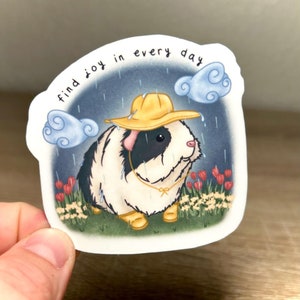 May include: A sticker featuring a cartoon guinea pig wearing a yellow raincoat and walking in the rain. The text "find joy in every day" is written in a handwritten font above the guinea pig.
