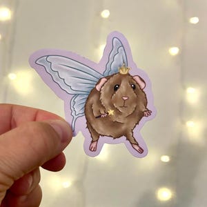May include: A sticker featuring a brown guinea pig with fairy wings and a gold crown. The guinea pig holds a magic wand. The sticker has a light purple border and is held by a hand against a background of fairy lights.