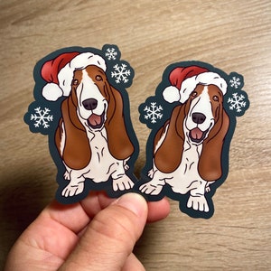 Puede incluir: Dos perros basset hound marrones y blancos con gorros de Papá Noel rojos con copos de nieve en el fondo.