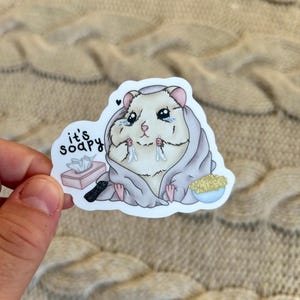 May include: A sticker featuring a cartoon hamster wrapped in a blanket, crying, with a box of tissues, a remote, and a bowl of popcorn. The text "it's soapy" is also on the sticker. The sticker is held by a hand.