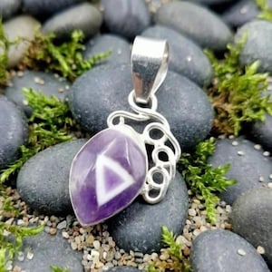 Pode incluir: Pingente de prata com uma pedra preciosa roxa com um design de triângulo branco. O pingente tem uma argola decorativa de prata e é colocado sobre um fundo de pedras cinzentas e musgo verde.