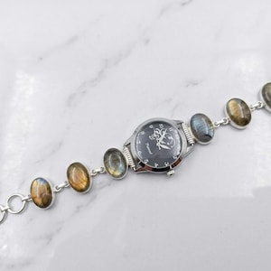 Op de afbeelding: Zilverkleurig horloge met een zwarte wijzerplaat en een decoratief ontwerp. De horlogeband heeft ovale, iriserende edelstenen. Het horloge ligt op een witte marmeren achtergrond.