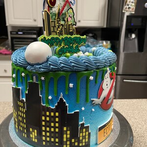 May include: A blue and green Ghostbusters themed birthday cake with a cityscape silhouette, green slime dripping down the sides, and a Ghostbusters logo on the side. The cake is decorated with white sprinkles and a Ghostbusters character topper.