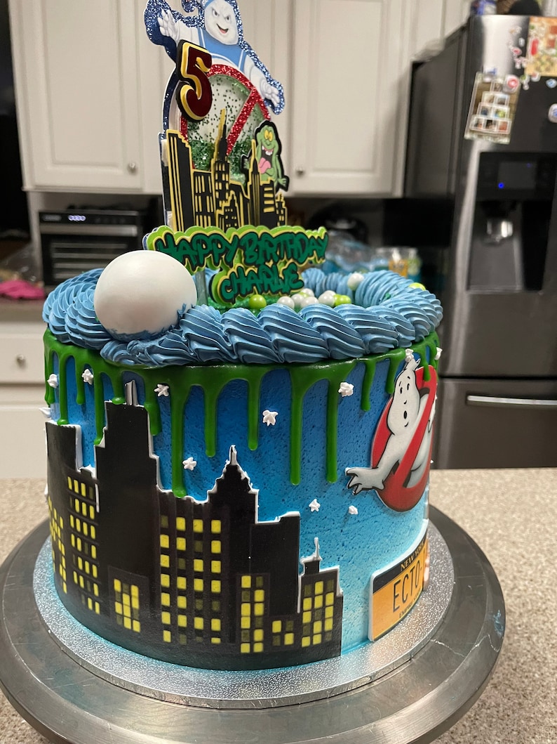 May include: A blue and green Ghostbusters themed birthday cake with a cityscape silhouette, green slime dripping down the sides, and a Ghostbusters logo on the side. The cake is decorated with white sprinkles and a Ghostbusters character topper.