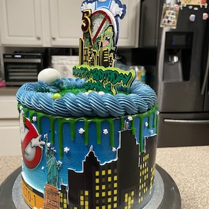 May include: A blue and green birthday cake with a Ghostbusters theme. The cake is decorated with a cityscape, a Statue of Liberty, and a Ghostbusters logo. The cake topper features a Ghostbuster character, a green monster, and the words "Happy Birthday Charlie".