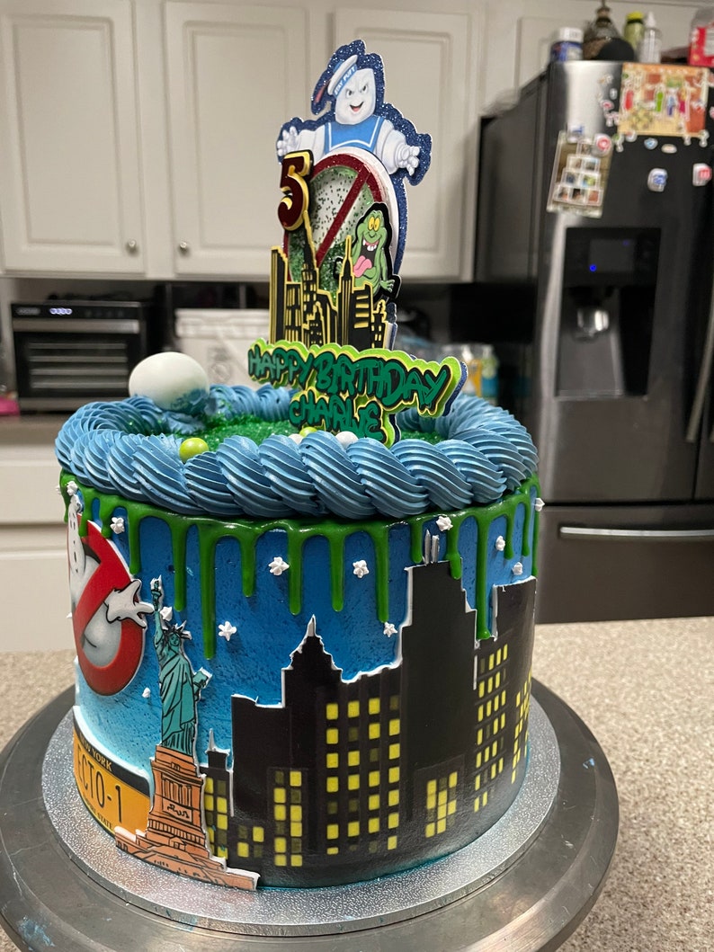 May include: A blue and green birthday cake with a Ghostbusters theme. The cake is decorated with a cityscape, a Statue of Liberty, and a Ghostbusters logo. The cake topper features a Ghostbuster character, a green monster, and the words "Happy Birthday Charlie".