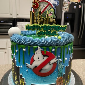 May include: A blue and green Ghostbusters themed birthday cake with a green slime drip, a ghost figure, and a New York license plate that says "ECTO-1".