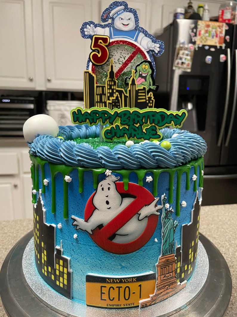 May include: A blue and green Ghostbusters themed birthday cake with a green slime drip, a ghost figure, and a New York license plate that says "ECTO-1".