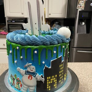 May include: A blue and green Ghostbusters themed cake with a white fondant ghost figure on top. The cake is decorated with green icing and white stars. The cake is on a silver cake stand.