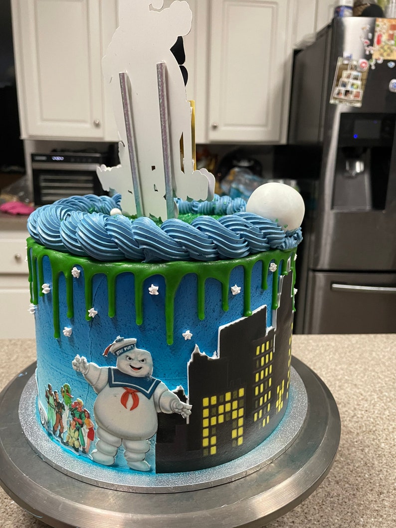 May include: A blue and green Ghostbusters themed cake with a white fondant ghost figure on top. The cake is decorated with green icing and white stars. The cake is on a silver cake stand.