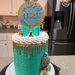 May include: A teal birthday cake with gold accents and a bow. The cake is decorated with a gold pattern and gold dripping icing. A topper reads "Happy Harlee 13".