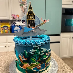 May include: A blue and green cake decorated with a Peter Pan theme. The cake is topped with a figurine of Peter Pan flying with Tinkerbell, a clock tower, and a moon. The cake is surrounded by a white frosting border.