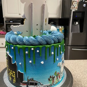 May include: A blue and green Ghostbusters themed cake with a white ghost figure on top. The cake is decorated with a cityscape, the Ghostbusters car, and green icing drips.