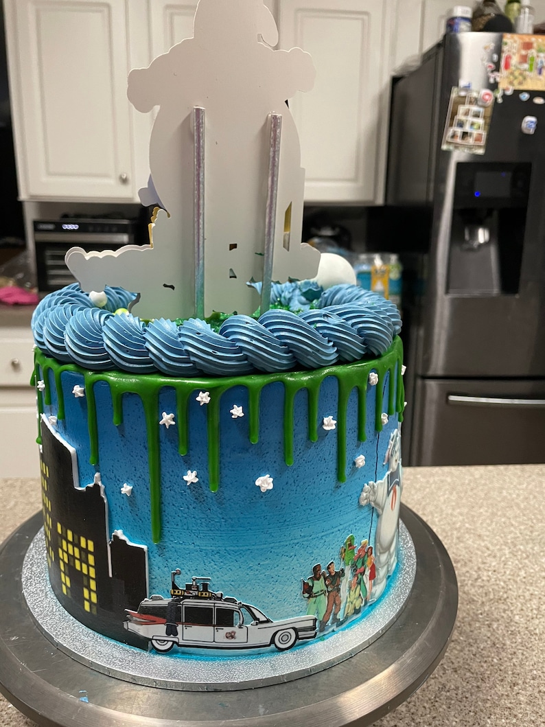 May include: A blue and green Ghostbusters themed cake with a white ghost figure on top. The cake is decorated with a cityscape, the Ghostbusters car, and green icing drips.