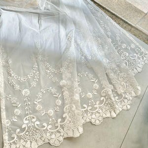 May include: A sheer white fabric with intricate floral lace detailing. The lace features a scalloped edge and delicate patterns of flowers and vines. The fabric appears to be lightweight and flowing, with a subtle sheen.