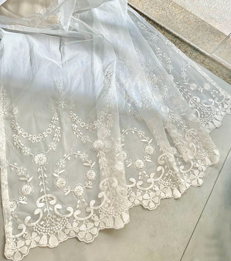 May include: A sheer white fabric with intricate floral lace detailing. The lace features a scalloped edge and delicate patterns of flowers and vines. The fabric appears to be lightweight and flowing, with a subtle sheen.