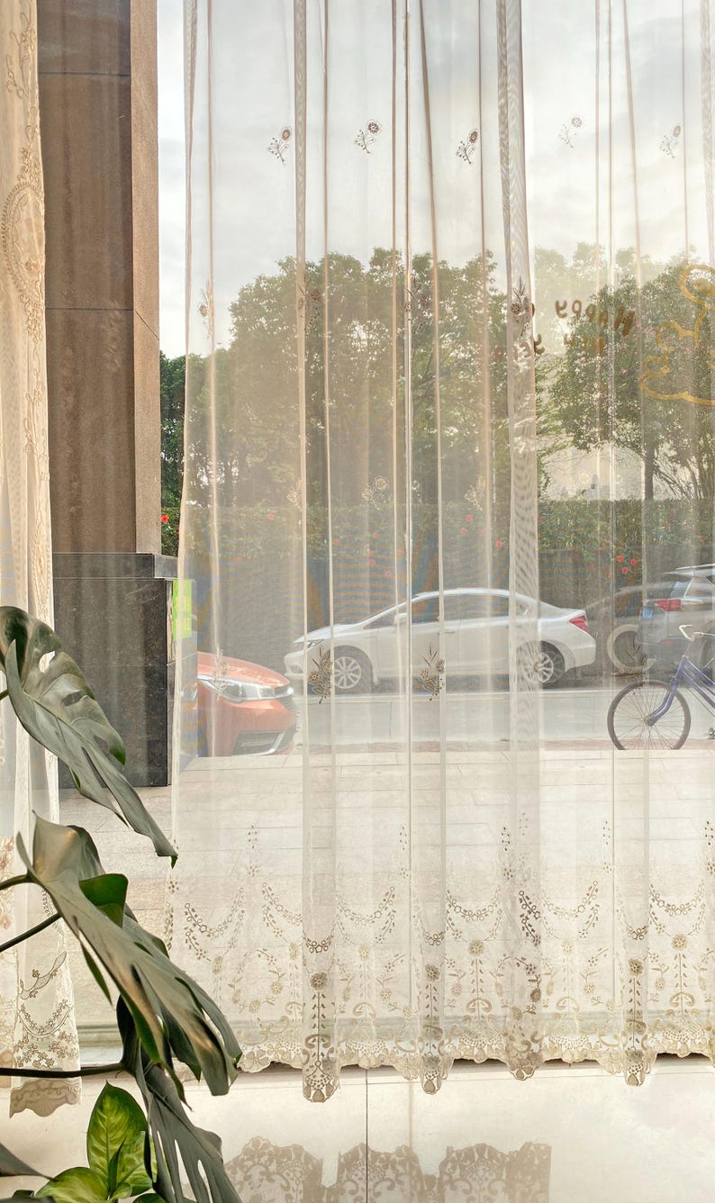 May include: Sheer, cream-colored curtains with delicate floral embroidery. The curtains are hanging in front of a window, with a view of trees, cars, and a bicycle outside. The light filters through the fabric, creating a soft, diffused glow.