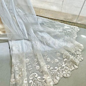 May include: White lace fabric with floral and leaf patterns. The delicate material is draped, showcasing the intricate embroidery along the edges. The fabric is likely used for decorative purposes, such as curtains or clothing.