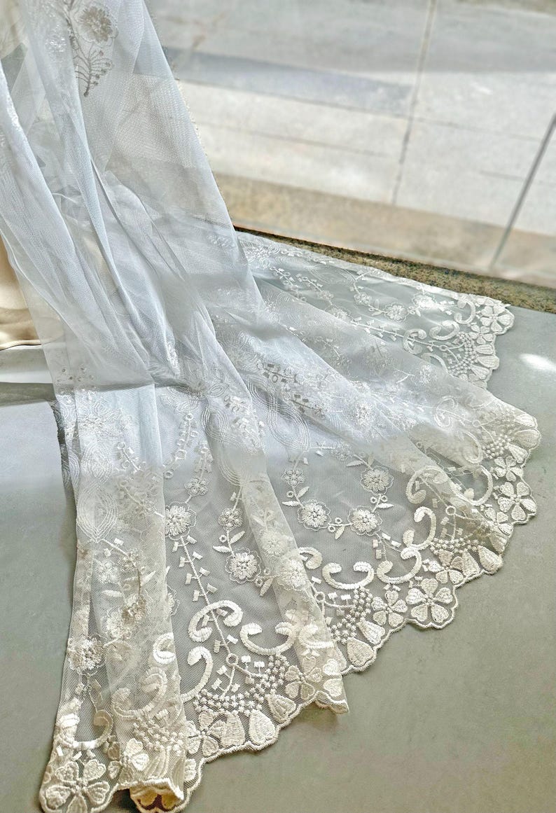 May include: White lace fabric with floral and leaf patterns. The delicate material is draped, showcasing the intricate embroidery along the edges. The fabric is likely used for decorative purposes, such as curtains or clothing.