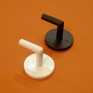 Può includere: Due ganci da parete, uno nero e uno bianco, con una base arrotondata e un braccio curvo. I ganci sono montati su una superficie arancione solida.