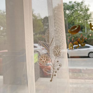 May include: Sheer, off-white curtain with a delicate floral embroidery in shades of beige and gold. The curtain's mesh-like texture allows light to filter through, creating a soft, diffused glow. The embroidered design features stylized flowers and leaves.