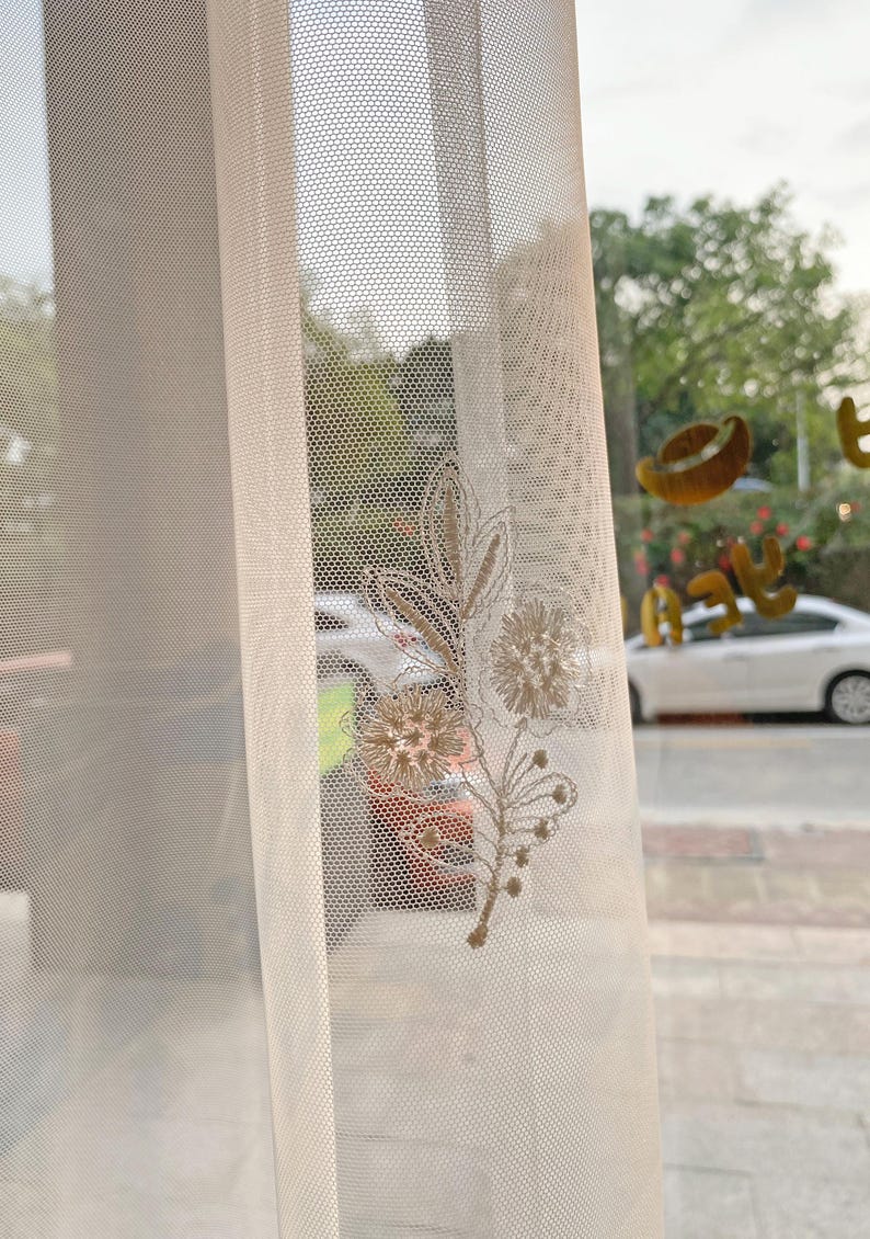 May include: Sheer, off-white curtain with a delicate floral embroidery in shades of beige and gold. The curtain's mesh-like texture allows light to filter through, creating a soft, diffused glow. The embroidered design features stylized flowers and leaves.